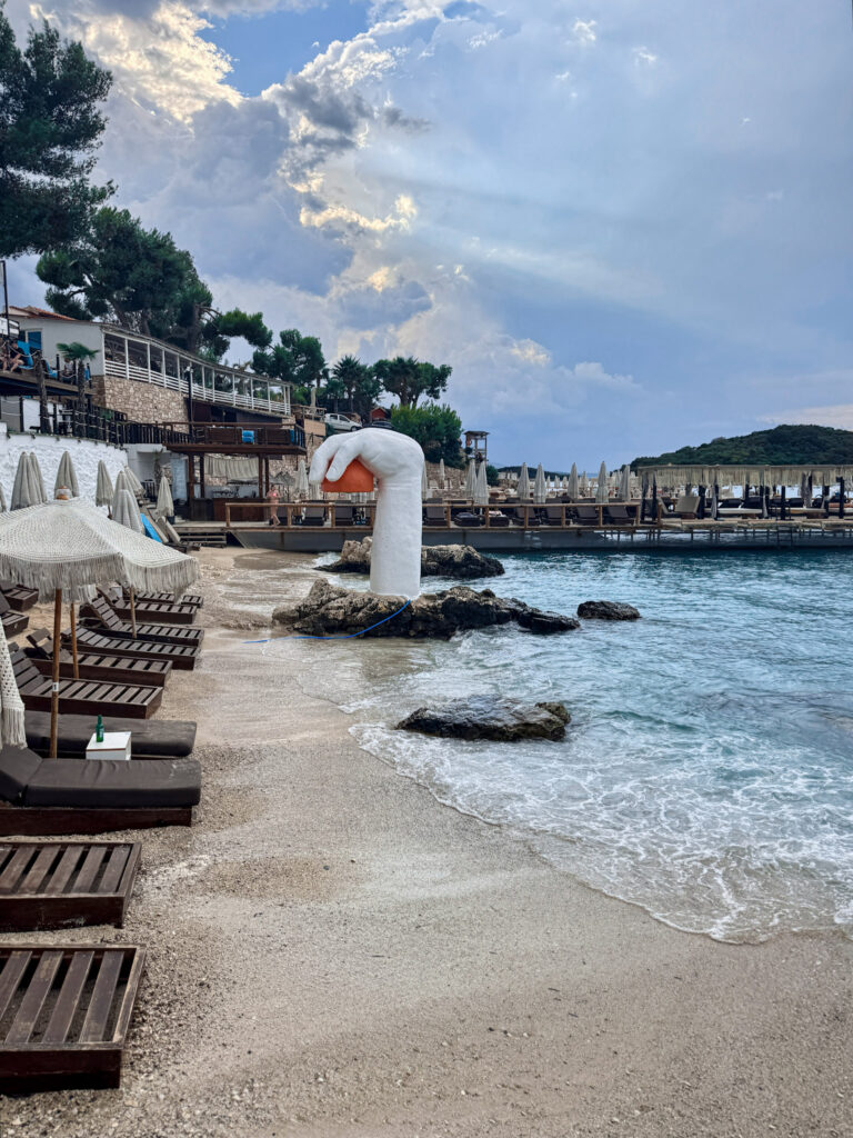 Top 10 Beaches in Ksamil, Albania The modern white hand sculpture rising out of the sea by rows of wooden sunbeds and fringed umbrellas at Plazhi Ksamilit in Albania.