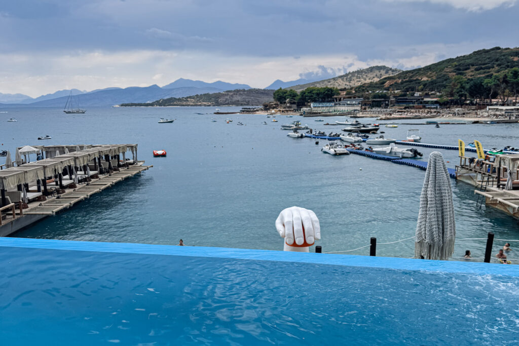 Top 10 Beaches in Ksamil, Albania Infinity pool view at Orange Beach Club in Ksamil, Albania, overlooking an overwater hand sculpture and a busy bay filled with boats.