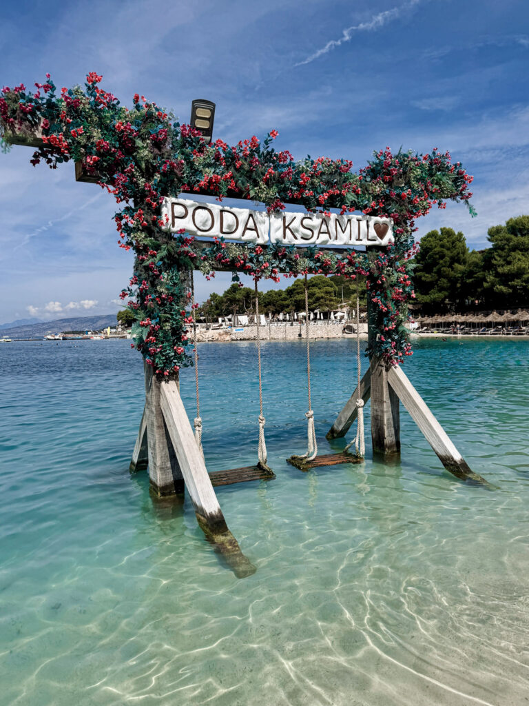 Top 10 Beaches in Ksamil, Albania The iconic Poda Ksamil ocean swing draped in pink flowers over crystal-clear turquoise water in Ksamil, Albania.