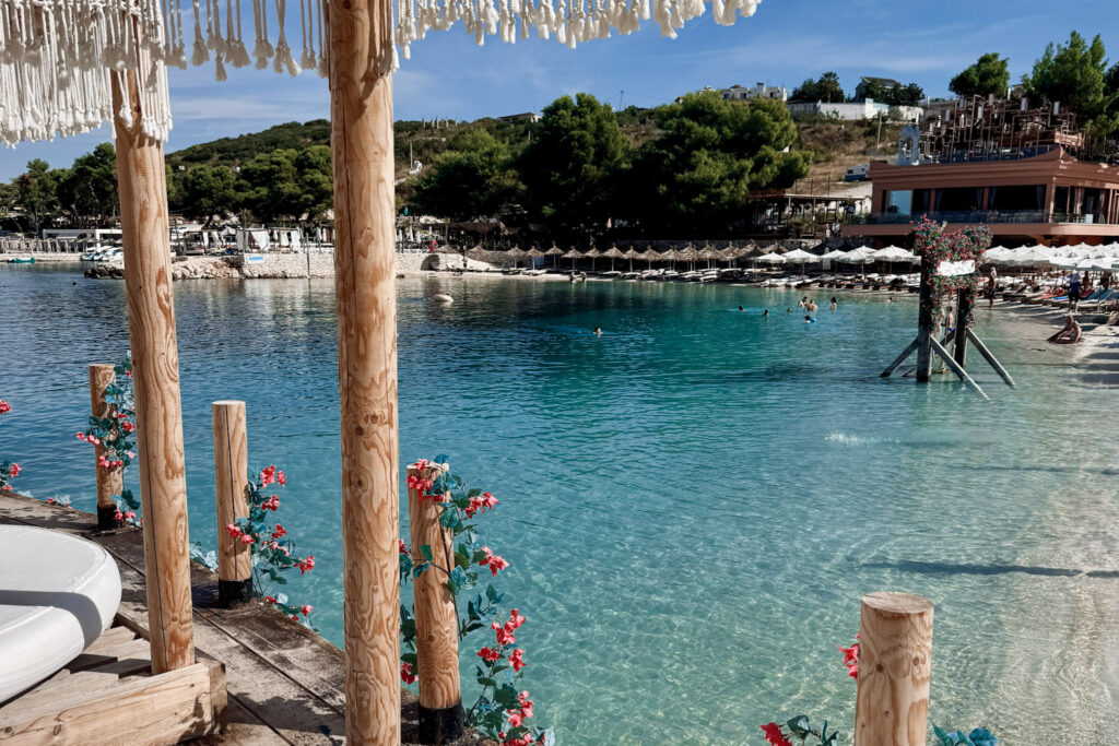 Top 10 Beaches in Ksamil, Albania View of a turquoise ocean cove from a wooden pier decorated with red flowers at Poda Beach Club in Ksamil, Albania.