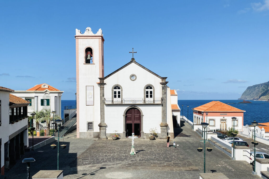 Madeira Travel Guide The historic Church of the Good Lord Jesus (Igreja do Senhor Bom Jesus) featuring a white facade and tall bell tower, situated in a cobblestone plaza by the sea in the coastal village of Ponta Delgada, Madeira.