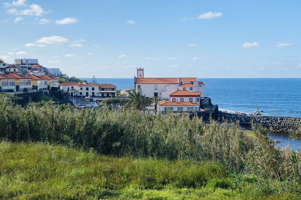 Madeira Travel Guide A scenic overview of Ponta Delgada, Madeira, showcasing the prominent white Church of Bom Jesus and orange-roofed coastal buildings nestled along a rocky shoreline next to the deep blue sea.