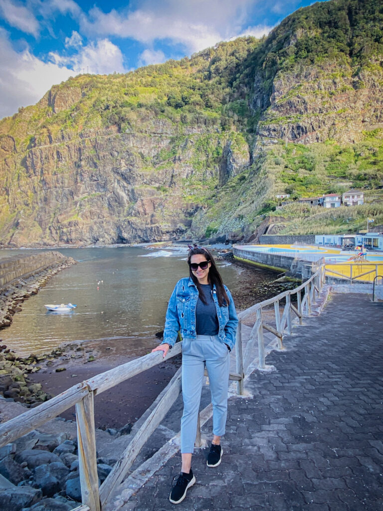Madeira Travel Guide A traveler posing on the coastal boardwalk next to the complex pools in Ponta Delgada, Madeira, with a quiet harbor cove and towering, lush green volcanic mountains rising in the background.