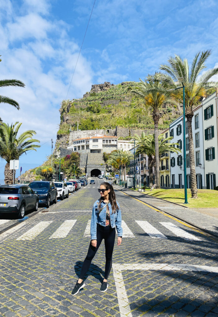 Madeira Travel Guide A traveler posing on a sunny cobblestone street lined with palm trees and cars, looking toward a large green hill and tunnel entrance in Ponta do Sol.