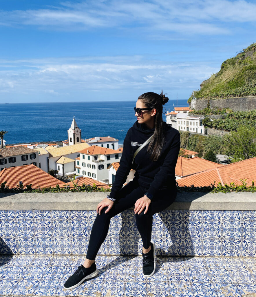 Madeira Travel Guide A traveler posing on a traditional Portuguese tiled wall at a scenic overlook, with a coastal village and the blue Atlantic Ocean in the background.