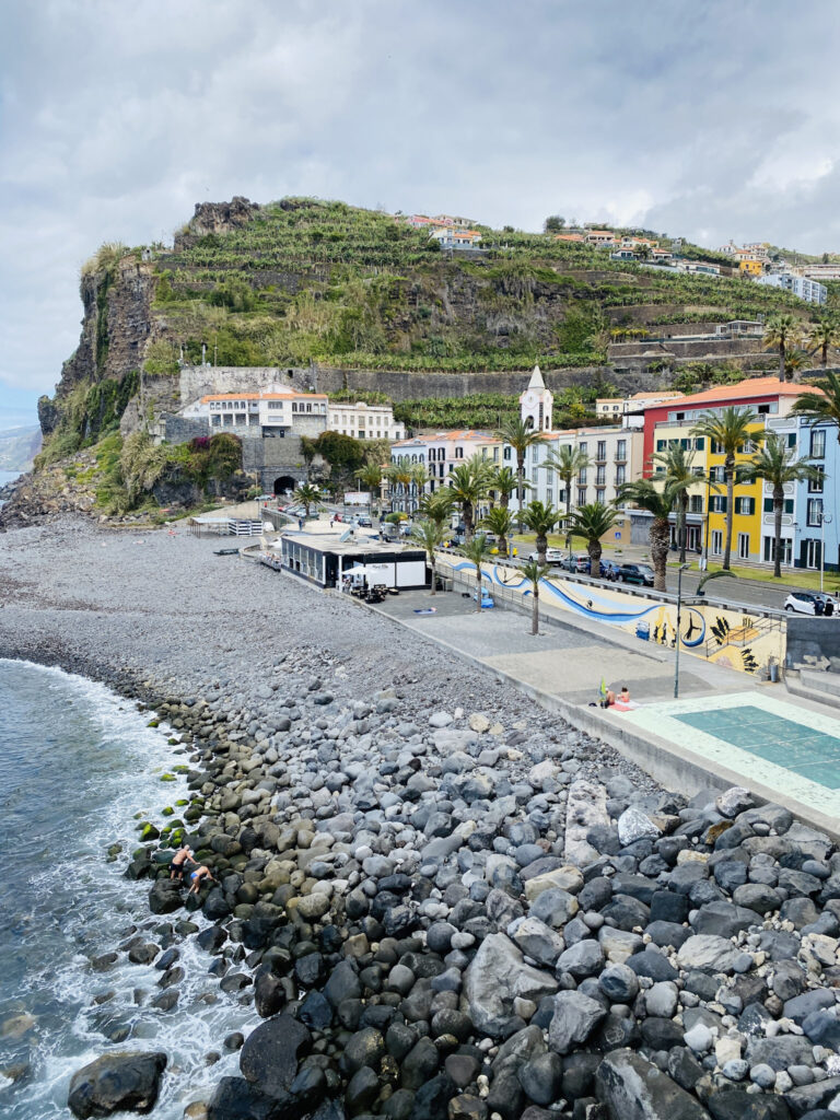 Madeira Travel Guide Elevated view of the rocky pebble beach, promenade, and colorful waterfront buildings of Ponta do Sol, Madeira under an overcast sky.