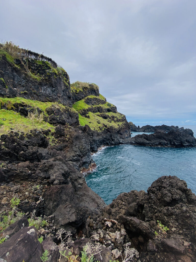 Madeira Travel Guide Rugged volcanic rock cliffs covered with green grass patches next to a blue ocean cove in Porto Moniz, Madeira.