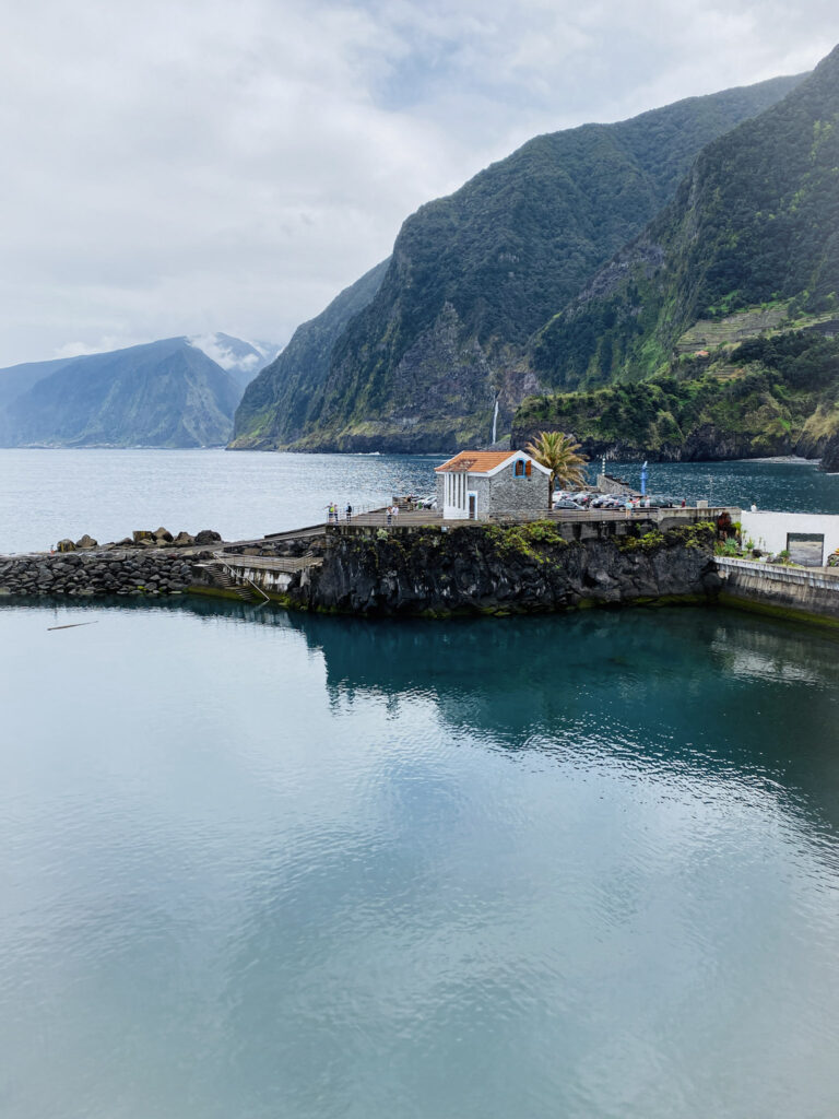 Madeira Travel Guide A small stone building with an orange roof on a pier overlooking a calm blue bay and towering green coastal mountains in Madeira.