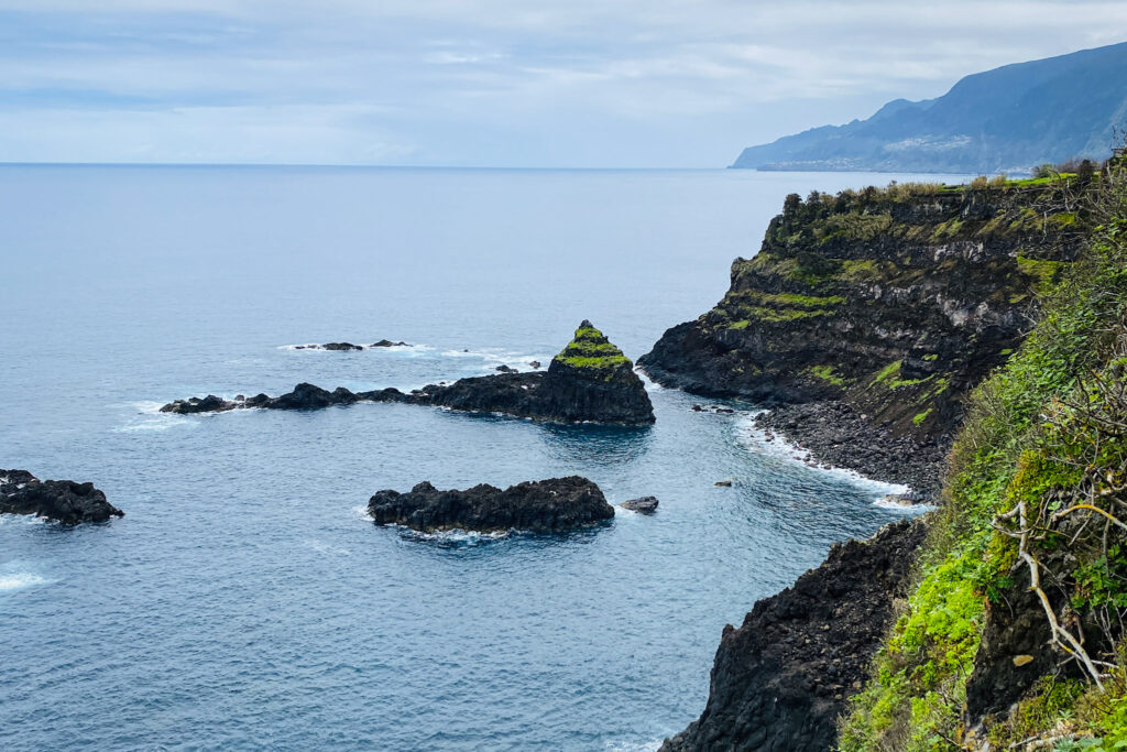 Madeira Travel Guide High-angle view of rugged volcanic cliffs and small rocky sea stacks topped with green grass in the blue ocean along the Madeira coast.