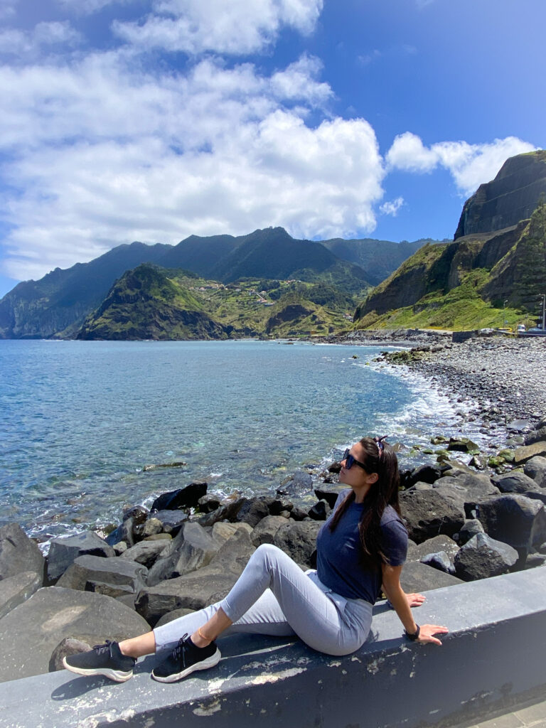 Madeira Travel Guide A traveler posing on a concrete barrier above a rocky black lava shore, with an expansive background of a calm blue ocean bay and towering, lush green mountains along the Porto da Cruz coastline in Madeira.