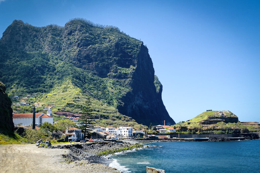 Madeira Travel Guide Scenic coastal view of the village of Porto da Cruz in Madeira, featuring white orange-roofed houses spread across green terraced fields under the imposing shadow of the majestic Penha de Águia mountain cliff next to a calm blue sea.