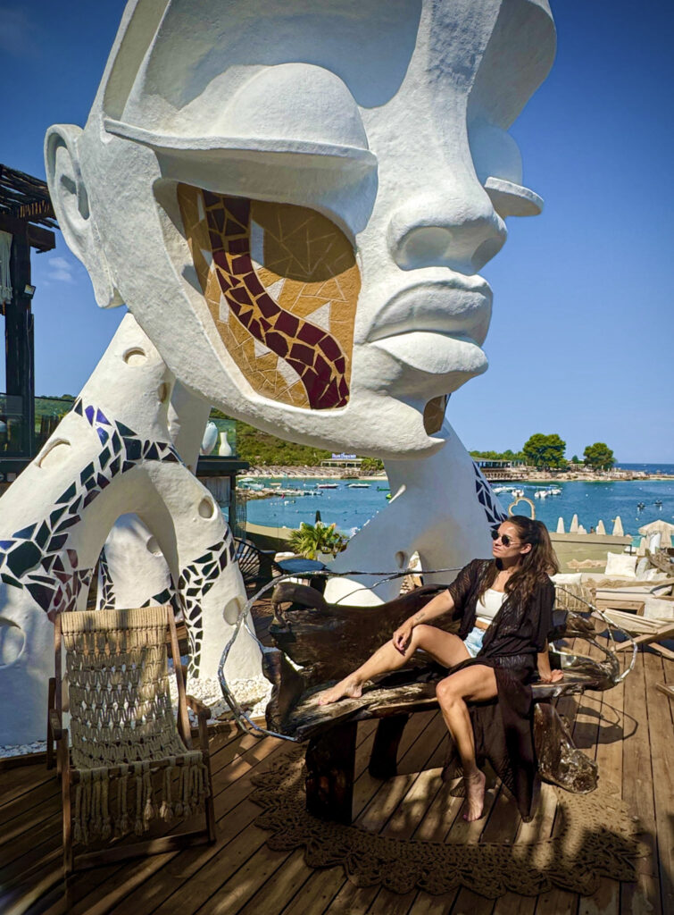 Top 10 Beaches in Ksamil, Albania Blog author posing on a rustic wooden bench next to a giant white bohemian head sculpture at Principotes Beach Club in Ksamil, Albania.