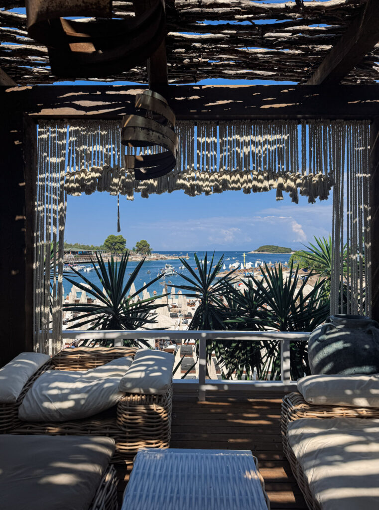 Top 10 Beaches in Ksamil, Albania View of the turquoise ocean bay and sunbeds framed by bohemian macrame curtains from a luxury terrace at Principotes Beach Club in Ksamil, Albania.