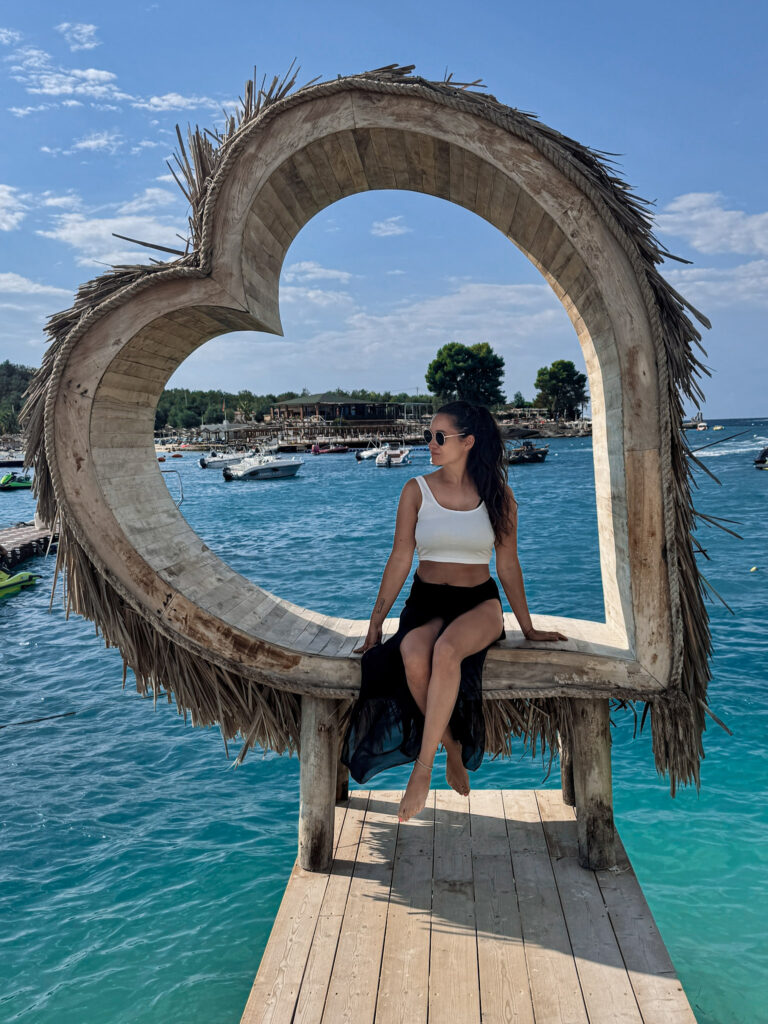 Top 10 Beaches in Ksamil, Albania Blog author sitting inside a large wooden heart-shaped frame on a pier overlooking the turquoise sea at Principotes Beach Club in Ksamil, Albania.