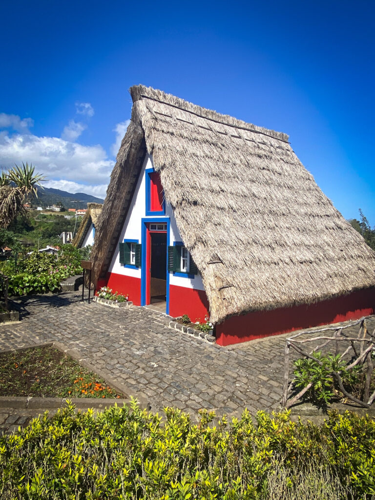 Madeira Travel Guide A traditional triangular thatched-roof cottage painted in white, red, and blue on a cobblestone courtyard in Santana, Madeira.