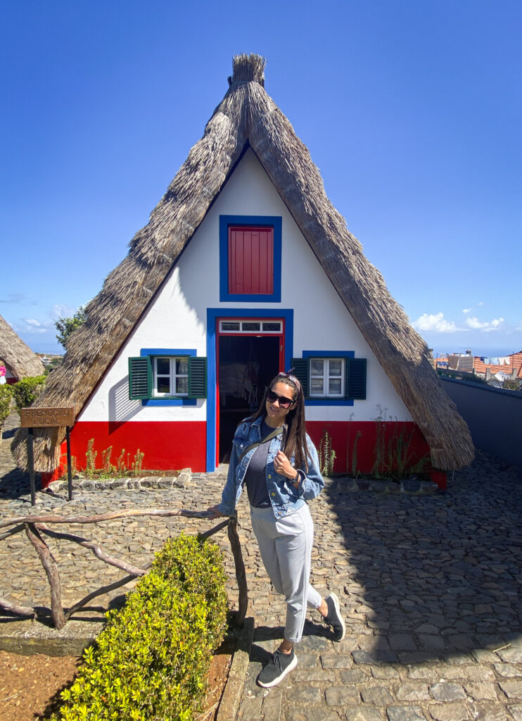 Madeira Travel Guide A traveler posing on a stone courtyard in front of an iconic, traditional Santana cottage featuring its unique triangular shape, thatched straw roof, and vibrant red, white, and blue paint in Santana, Madeira.