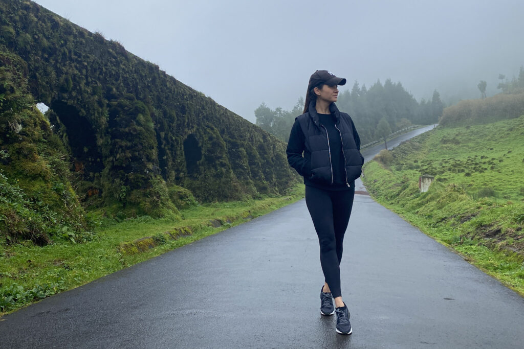Top Things to Do in São Miguel, Azores A traveler walking down a wet asphalt road next to the historic, moss-covered Muro das Nove Janelas aqueduct on a foggy day in São Miguel, Azores.