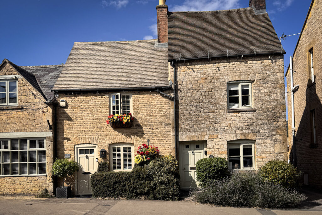 Top 5 Villages in the Cotswolds A row of traditional honey-colored Cotswold stone cottages on a street in Stow-on-the-Wold, England. The historic buildings feature tiled gabled roofs, cream-colored windows and doors, small green bushes, and wooden window boxes filled with bright red and yellow flowers under a clear, bright blue sky.