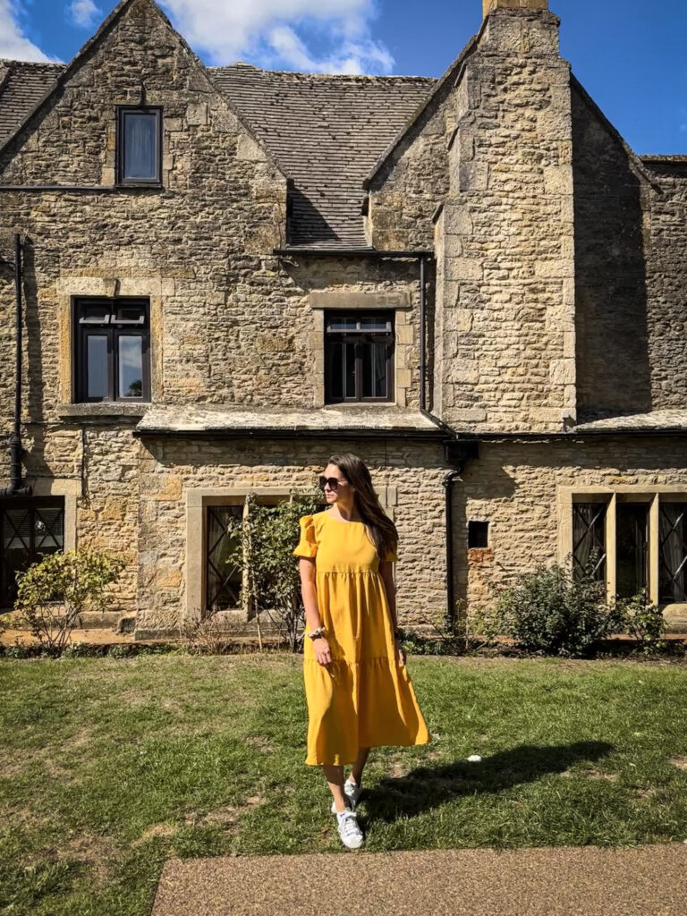 Top 5 Villages in the Cotswolds Standing on a sunny green lawn in front of a historic honey-colored Cotswold stone building. The sun casts her shadow onto the grass, while the rustic limestone facade features traditional multi-paned glass windows and leaded window frames.
