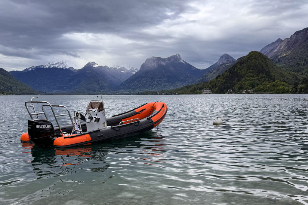 Top 10 Places in Southeastern France An orange and black inflatable rigid hull boat floats on the calm green water of Lake Annecy, with lush green hills and dramatic mountains under a moody, overcast sky in France