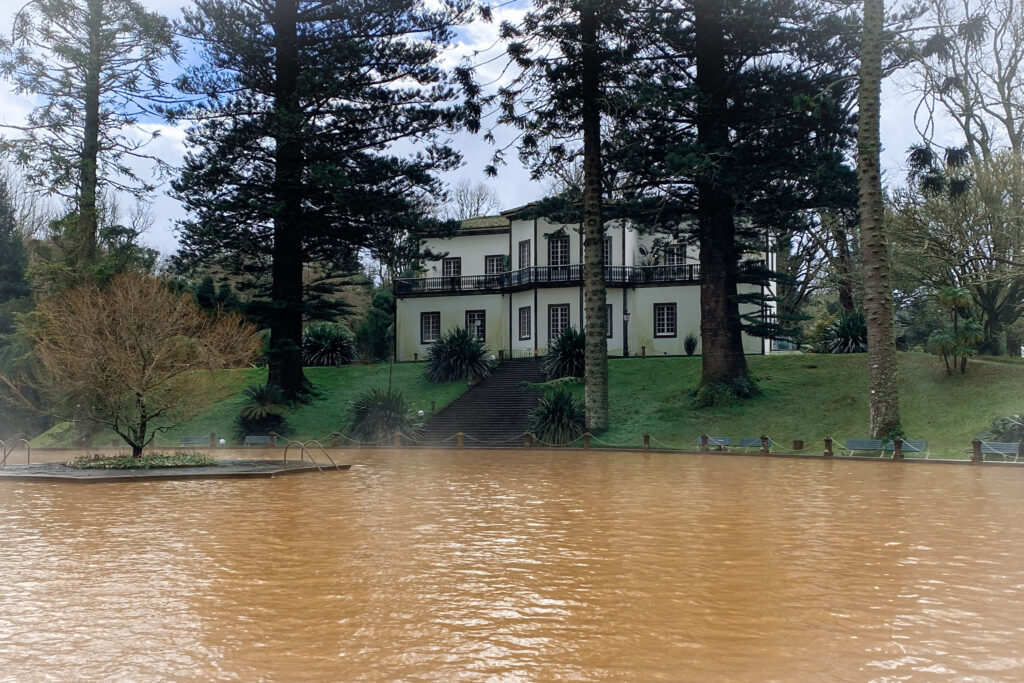 Top Things to Do in São Miguel, Azores The large, muddy-orange iron-rich thermal swimming pool in front of the historic white Terra Nostra Garden Hotel mansion framed by tall pine trees in Furnas, São Miguel, Azores.