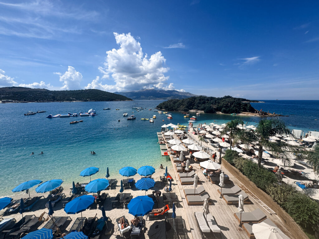 Top 10 Beaches in Ksamil, Albania Elevated view of Three Islands Beach in Ksamil, Albania, showing a turquoise bay filled with boats, white sunbeds, and lush green islands.