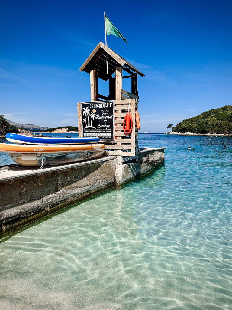Top 10 Beaches in Ksamil, Albania A wooden lifeguard tower with a green flag and stacked paddleboards on a concrete pier over clear turquoise water at Tre Ishujt Restaurant & Lounge in Ksamil, Albania.