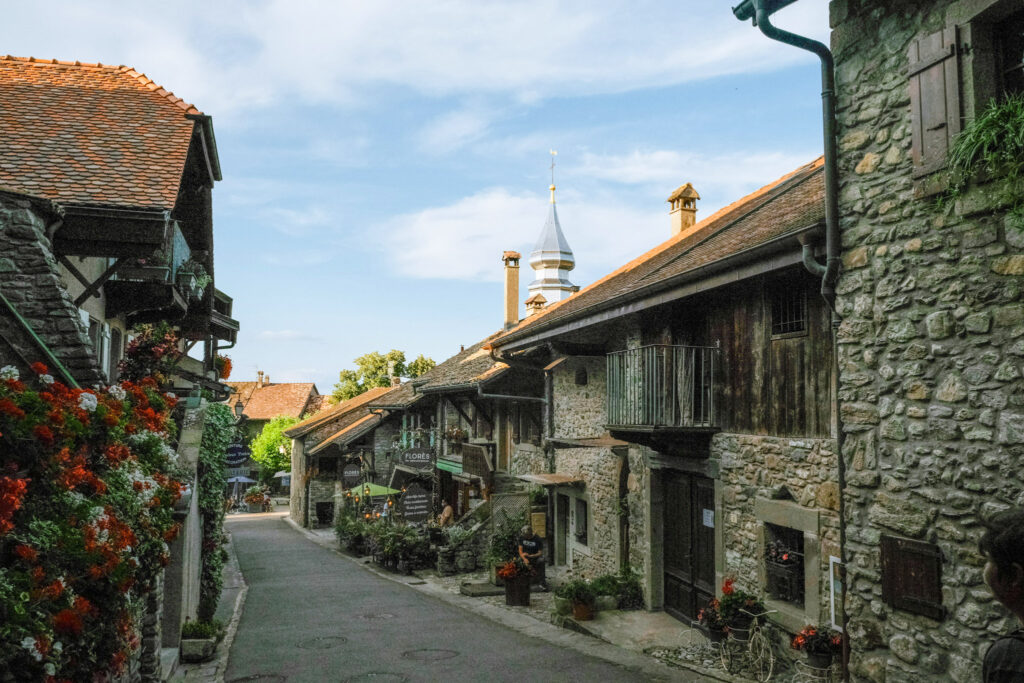Top 10 Places in Southeastern France A narrow, sloped street lined with traditional stone and wood-trimmed houses decorated with cascading flowers, leading toward the distinct silver onion dome steeple of Églises Saint Pancrace under a blue sky in the medieval village of Yvoire, France