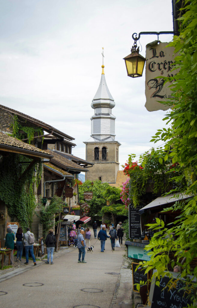 Top 10 Places in Southeastern France Tourists stroll down a narrow pedestrian street lined with ivy-covered stone houses and artisan shop signs, leading toward the prominent silver onion dome steeple of the Saint-Pancrace church under an overcast sky in Yvoire, France
